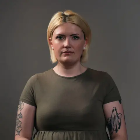 BBC Scotland Robyn, who has a blonde bob, is staring straight at the camera. She is wearing a khaki green dress and has tattoo on her arms. 