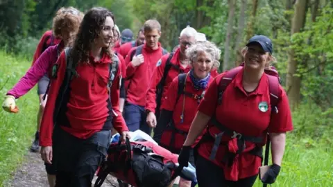 Multiple people walking through a forest in red matching uniforms. People at the front of the group are carrying a stretcher, which appears to be filled with equipment.