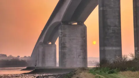 MrBlueSky/BBC Weather Watchers A photo from below large concrete structure of the Orwell Bridge during a sunrise on a slightly misty morning. The River Orwell flows below the bridge. 