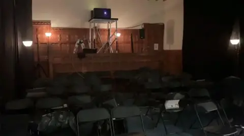 A rear-facing photo of a magistrates court being repurposed as a cinema. A projector sits on the high bench were the magistrates would have sat and rows of folding chairs are arranged on the floor below