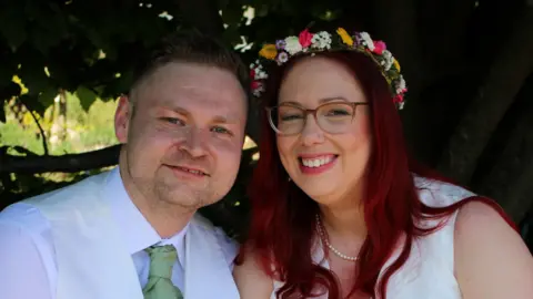 St Elizabeth Hospice A close-up of a man and a woman. The man is wearing a white shirt and green tie under a white waistcoat. The woman is wearing glasses, a pearl necklace, and a wedding dress. They are both smiling and looking into the camera.