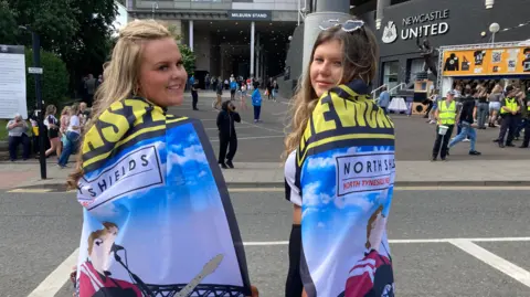 Two young women on their way to the Sam Fender concert. They are outside the Milburn Stand at St James' Park where it curves round towards the Gallowgate. The words Newcastle United, along with the club badge, are on the black brickwork behind a statue of Alan Shearer. People are gathered by a merchandise stall next to the statue. The two fans have their backs to the camera but have their heads turned to face it. They are wrapped in Sam Fender flags that include a cartoon of the singer and a street sign which reads North Shields, North Tyneside.