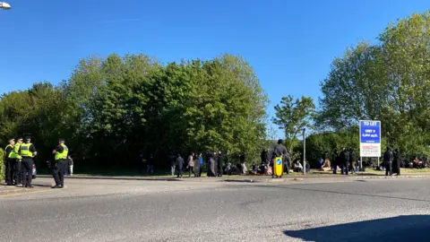 A group of police officers stand on a road, with a large group of people wearing dark clothing stood to the left, next to a "to let" sign. There are trees behind them all.