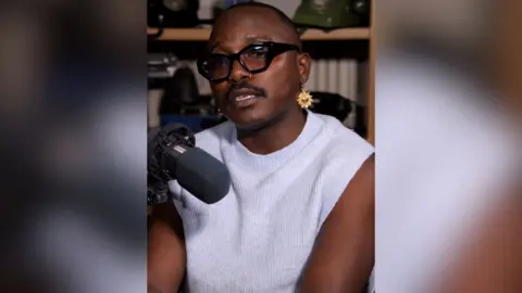 A man in a white vest top sits in front of a microphone with a bookcase behind him
