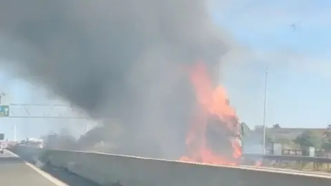 Flames and thick smoke rise from a lorry fire at the side of a major road.