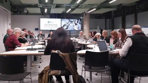 Nadia Lincoln/LDRS Councillors sitting around a table during a meeting. Many of them have laptops and microphones in front of them. Meeting papers are being shown on a large TV screen.