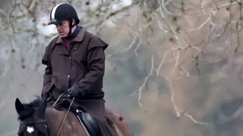 Reuters Andrew wearing a riding helmet as he rides a brown horse against a backdrop of tree branches in Windsor in February 2026