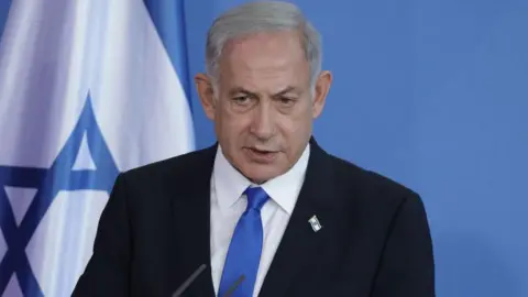 A shot of Netanyahu from the shoulders up, wearing a dark suit with a white shirt and a light blue tie. He has a small pin of the Israel flag on his lapel. Behind him is a plain blue background with part of the Israel flag to the left.