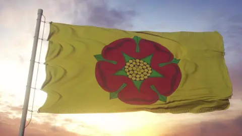 A large yellow flag with a red rose with green leaves and and yellow centre painted on it - it flies in front of a sunset