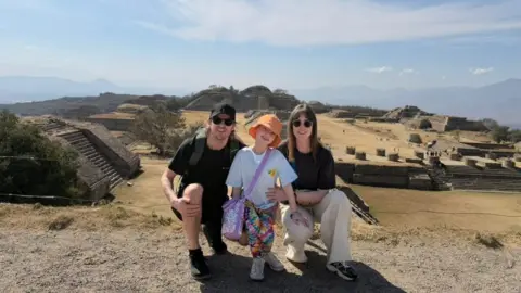 Marratravels The Henry family posing for a photo on front of an archaeological site featuring pyramid-shaped tombs. Craig and Alli are crouching down either side of Summer. Craig is wearing a black t-shirt and shorts with a black baseball cap and sunglasses. Summer is wearing a white t-shirt with tie-dye stile multicoloured leggings and orange bucket hat. Alli is wearing a black t-shirt, cream trousers and sunglasses.