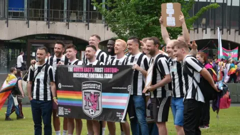 A group of a dozen Newcastle United fans, wearing the black and white strip, and holding a banner stating "The North East's First LGBTQIA+ Inclusive Football Team.