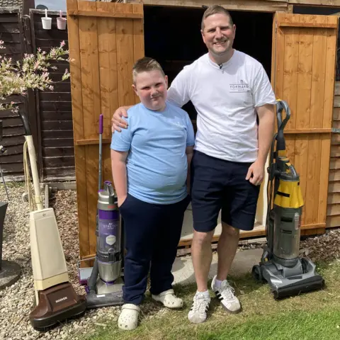 A boy and a man stood outside a garden shed smile at the camera. The man has one arm around the boy and the boy has his arm in his pocket. They're stood next to three standing vacuum cleaners. The boy is wearing navy blue jogging bottoms, a light blue T-shirt and white clogs. The man is wearing navy blue shorts, a white T-shirt and white trainers.