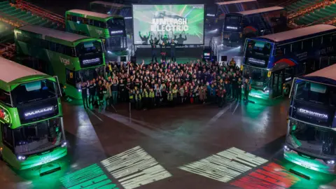 Eight green and blue double decker buses are parked up around the large arena with a crowd of people standing between then and cheering at the camera. A screen behind them reads: "Unleash electric".