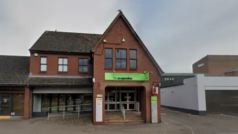 A Google street view image shows the front of the Co-Op on The Avenue. A two-storey red brick building, a green sign reading "The co-operative" sits above the entrance. Beneath it and to the right is a Post Office sign. A lane goes down the right hand side of the shop as we look at it.
