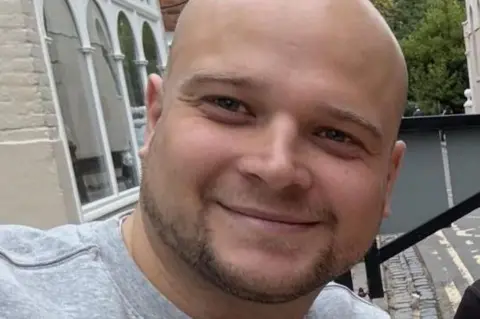 George Balkwill George smiles for the camera. He has a bald head and brown facial stubble. He wears a light grey shirt.