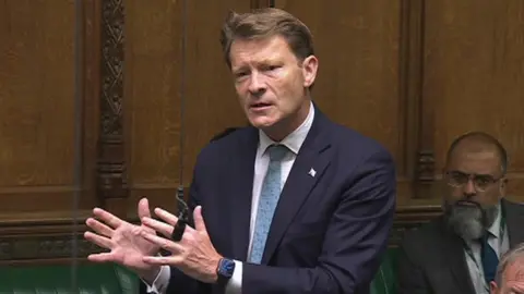 Richard Tice wearing a blue suit, with white shirt and light blue tie stood in parliament delivering his speech, with both hands pointing to his right as he makes his point heard