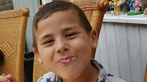 Family A boy sits on a wicker chair outside and smiles at the camera. He has dak brown hair and he is eating ice cream.