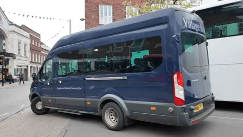 A dark blue mini bus that has four wheels and is parked on a grey road.