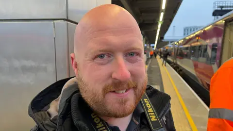 A man looks into the camera. He was an auburn beard. He's wearing a black coat and has a camera strap over his neck. He's standing on a train platform and smiling.