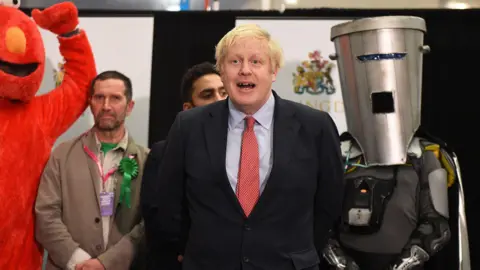 Getty Images Boris Johnson is in the centre of the photo, wearing a black suit with a pale blue shirt and a red spotted tie. Behind him to the right is Count Binface, wearing a large silver bin on his head and a silver cape. On his right is a man dressed as the fuzzy red Sesame Street character Elmo.