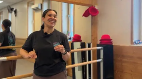 Maryam Pourian is pictured smiling looking to the right of the camera. She is stood in a dance studio with a large mirror behind her.