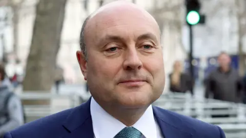 A man is standing in front of the camera but nt looking into the lense. He's wearing a navy suit with a white shirt and a tie. The background is blurred.