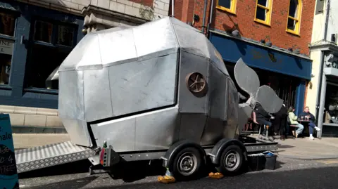 The whale looks sort of like a silver metal caravan that has been cut up and re-arranged into a whale shape.  A wheel has been used for it's eye.  It sits on a trailer.