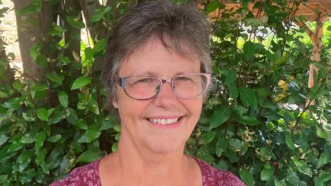 Shelley Porter. She has short grey hair and is wearing pink/silver glasses and a pink top. She is stood in front of a bush.