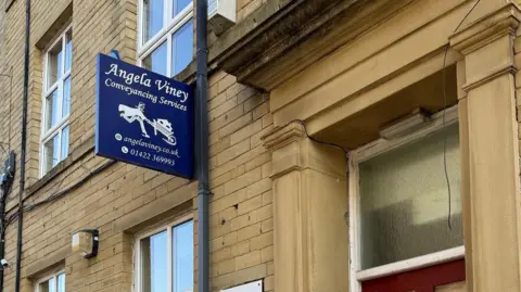 Angela Viney A blue hanging sign reads “Angela Viney Conveyancing Services” along with a stylised white horse logo and additional contact details. Various wall-mounted lights, alarms, and security devices are also visible on the building’s exterior.
