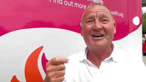 John, wearing a white shirt, standing in front of a pink and white clinical testing van.