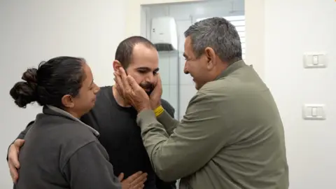 In a khaki jacket, a man keeps the face of Yardon Bibas as he hugs a relative after being released from Hamas's captivity. 