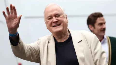 An elderly, grey haired gentleman in a white suit , waving at a crowd