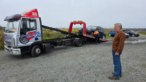 Andrew Cameron A large pick-up lorry is seen with a black car on the back while in the foreground stands a man with blue jeans and a brown jumper. 