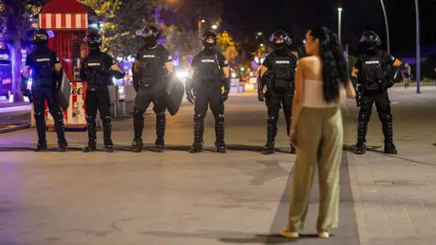 A woman in a white top and green trousers stands in front of riot police dressed in black
