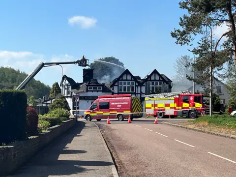 Jim Sneddon Firefighters use an extended aerial platform to spray water onto the roof of a damaged building in daylight, with a fire investigation van, fire engine and cordon visible outside the property.