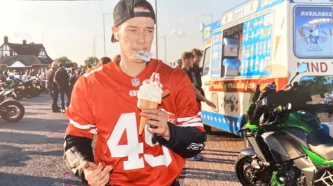 A young man wearing a padded biking jacket covered by a red American football shirt and a black baseball cap on backwards is walking past numerous motorbikes and an ice cream van. He is holding an ice cream in his hand and a ten pound note in his mouth.