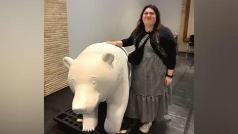 Becky Shepperson in a long dress and cardigan, smiling with her hand on a bear sculpture. The bear is white as it has not been painted yet.
