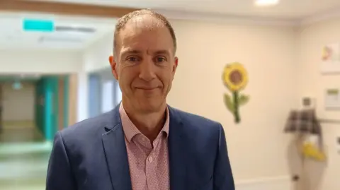 Kenny Steele standing inside the hospice. There are light yellow walls, one of whcih has a sunflower on it. He has short hair and is wearing a red patterned shirt and a blue suit jacket.