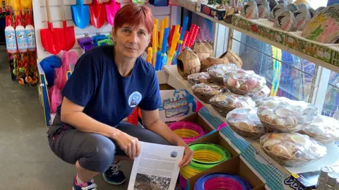 Clair Murgatroyd Clair Murgatroyd in a seaside shop crouching next to a display of plastic toys, she holds a printed newsletter with a photo of a dead seal. 