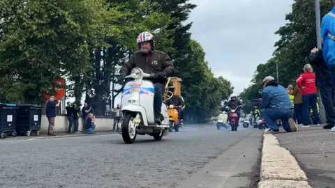 A man rides a cream Lambretta as pedestrians stop on the pavement to watch the parade of scooters pass by