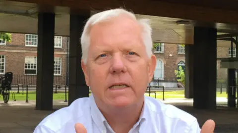 Daniel Mumby David Fothergill, a man with short white hair, looking at the camera. He is wearing a white shirt. Windows of a brown brick building can be seen behind him.