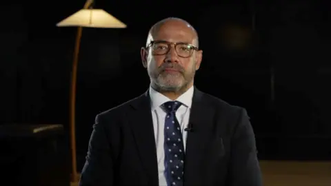 Neil Basu looks straight down the lens with a neutral expression, in a dark room. He is wearing dark-framed glasses, a black suit and a dark blue tie with white polka dots. 