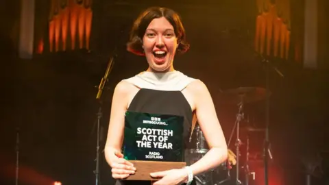 Alice Faye wearing a black and white dress holding the trophy for BBC Introducing Scottish Act of the Year. 