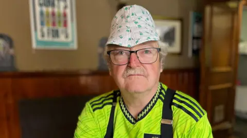 Dave Nelson is wearing a luminous yellow, green and black Manchester United away top with black braces. He has black-rimmed glasses, short white hair and a moustache. He is also wearing a small white hat with a design of little green flowers on it.