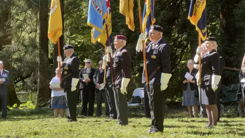 A group of around six people standing on some grass below tall trees. They are wearing military uniforms and holding up large flags during a memorial ceremony on a sunny day.