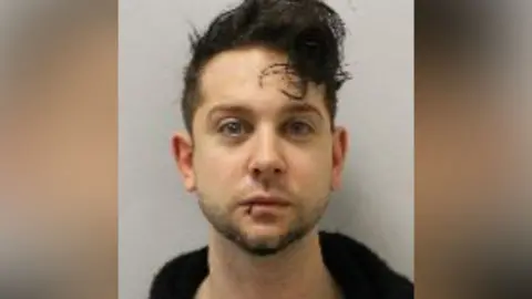 Met Police Mugshot of a man with dark hair and short beard. He looks directly at the camera and wears a black jumper.