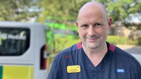 Ben Ford is standing in front of an ambulance wearing a blue NHS outfit. He is smiling at the camera