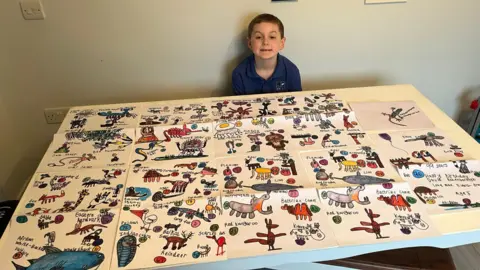 Max Evans-Browning Max Evans Browning sits behind a table which is covered in 18 pages of animal drawings. He smiles for the photo. 