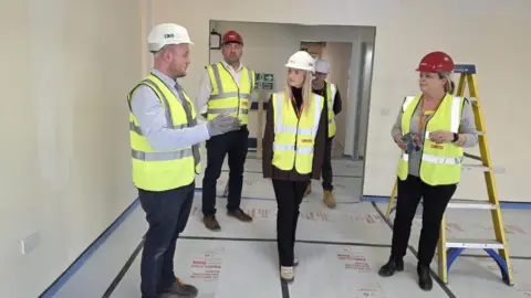 Office of Rosie Wrighting MP A blonde woman in a high visibility jacket and white hard hat walks in a room that is being refurbished. She is being spoken to by a man in a high visibility jacket and white hard hat. Several other people are in the room.