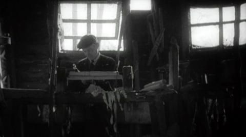 Black and white. A chair bodger sits inside, crafting a chair leg from a piece of wood using his tools.